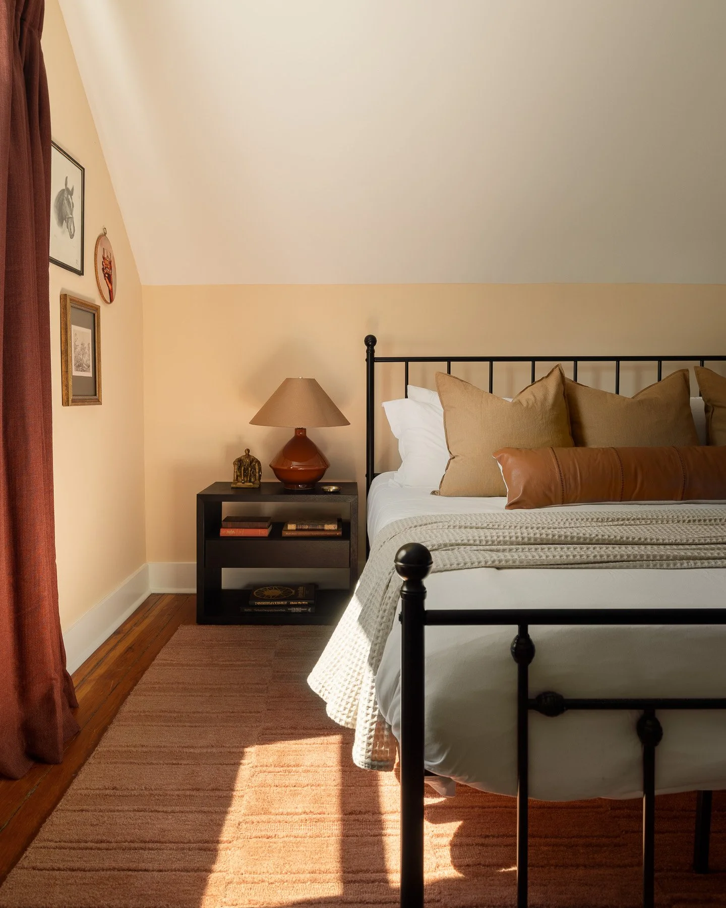 A bedroom corner with a black metal bed frame, beige pillows, a bedside table with a lamp, a small gold sculpture, and framed artwork on the beige wall. A window with a red curtain is partially visible on the left, with sunlight casting a shadow on the wooden floor.