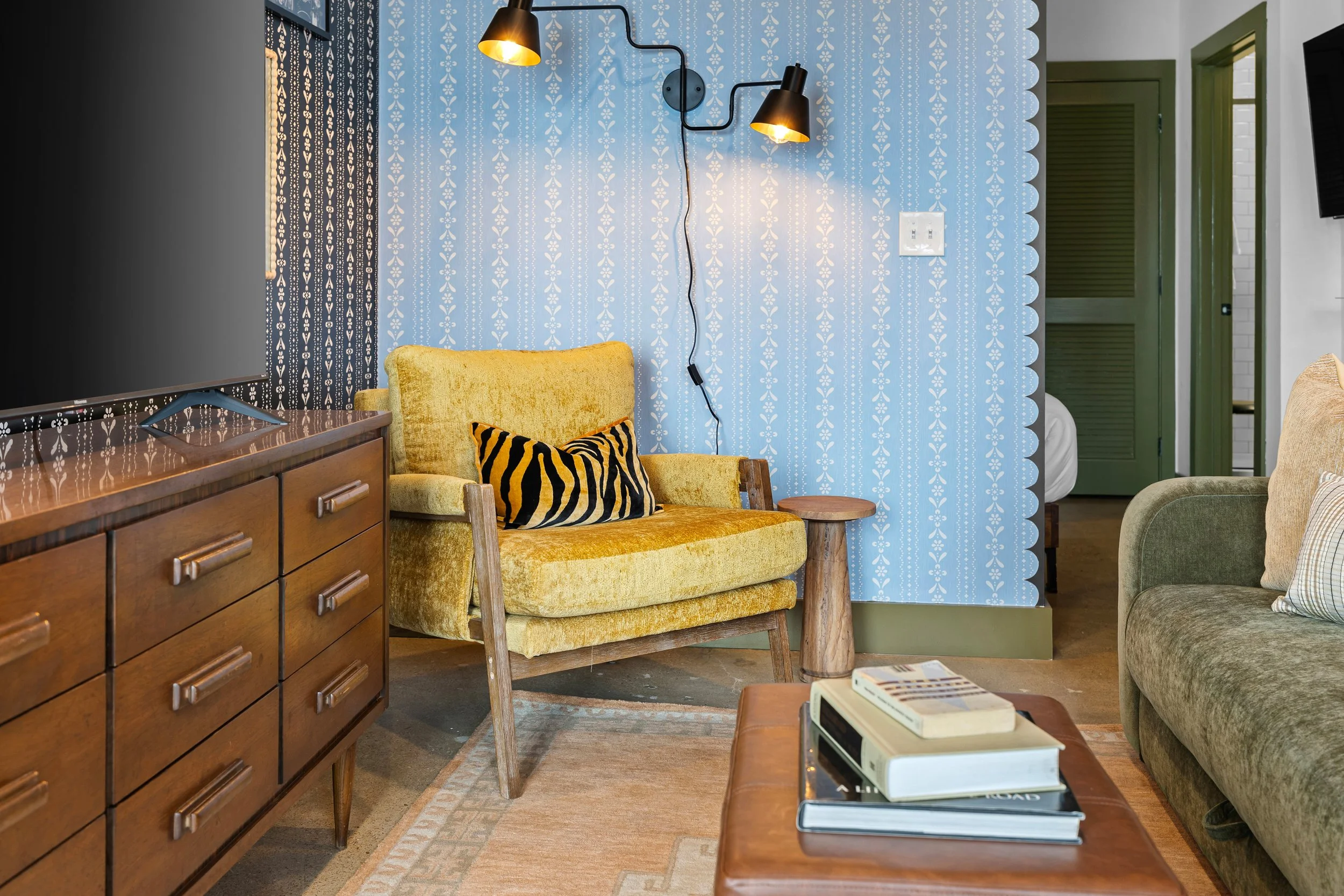 Living room with yellow armchair, wooden side table, and a coffee table with books. A distressed wood dresser with a TV on top is on the left. The wall behind the armchair has blue wallpaper with white floral patterns. Two black wall-mounted lamps with yellow interiors are on the blue wallpaper wall. To the right, a doorway with green trim leads to a bedroom with a visible white bed. The floor has a patterned rug.