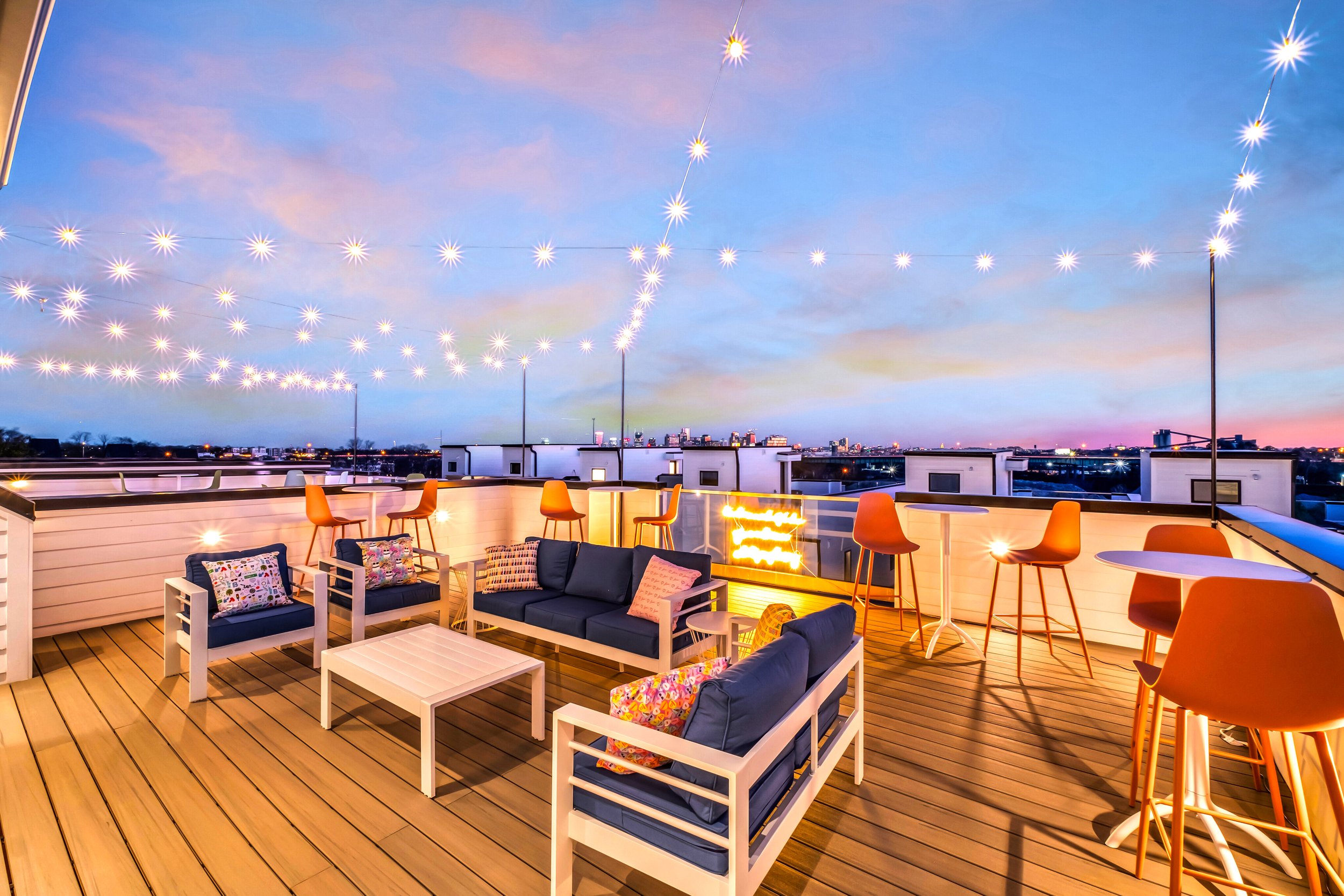 A rooftop patio during evening with string lights, modern outdoor furniture including sofas, chairs, and tables, overlooking a city skyline.