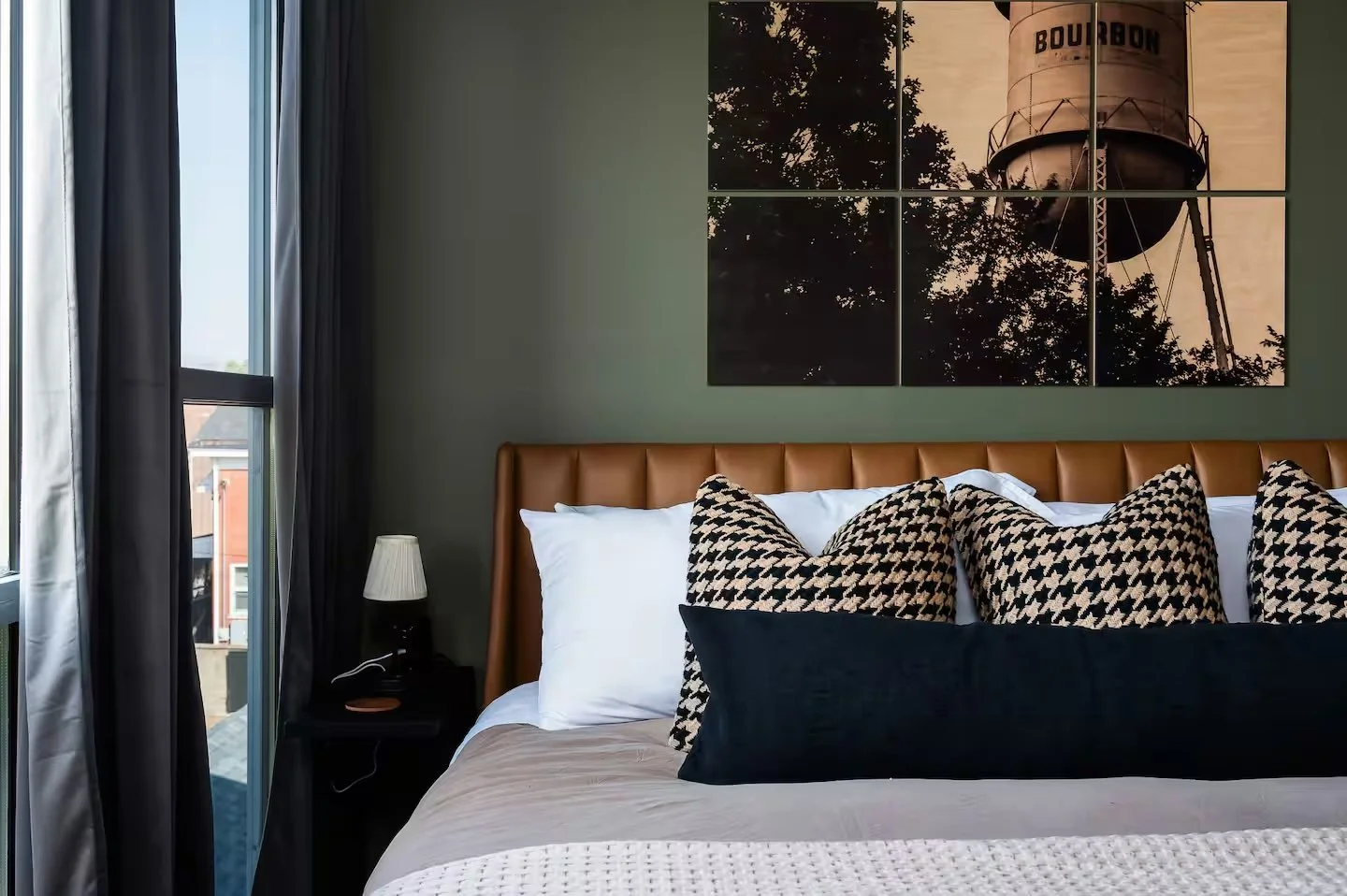 A bedroom with a bed featuring white and patterned pillows, a nightstand with a lamp, and a multi-panel artwork of a water tower on the green wall.