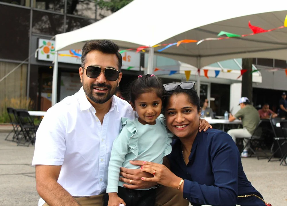 A mom and dad embracing their daughter outside during a family BBQ at Orange Seeds Montessori Centre