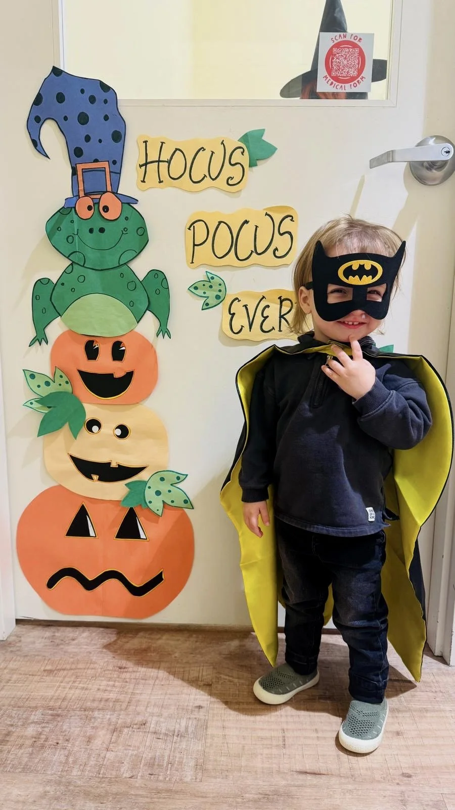 A little boy smiling with a batman Halloween costume on in front of paper cut-outs of jack-o-lanterns