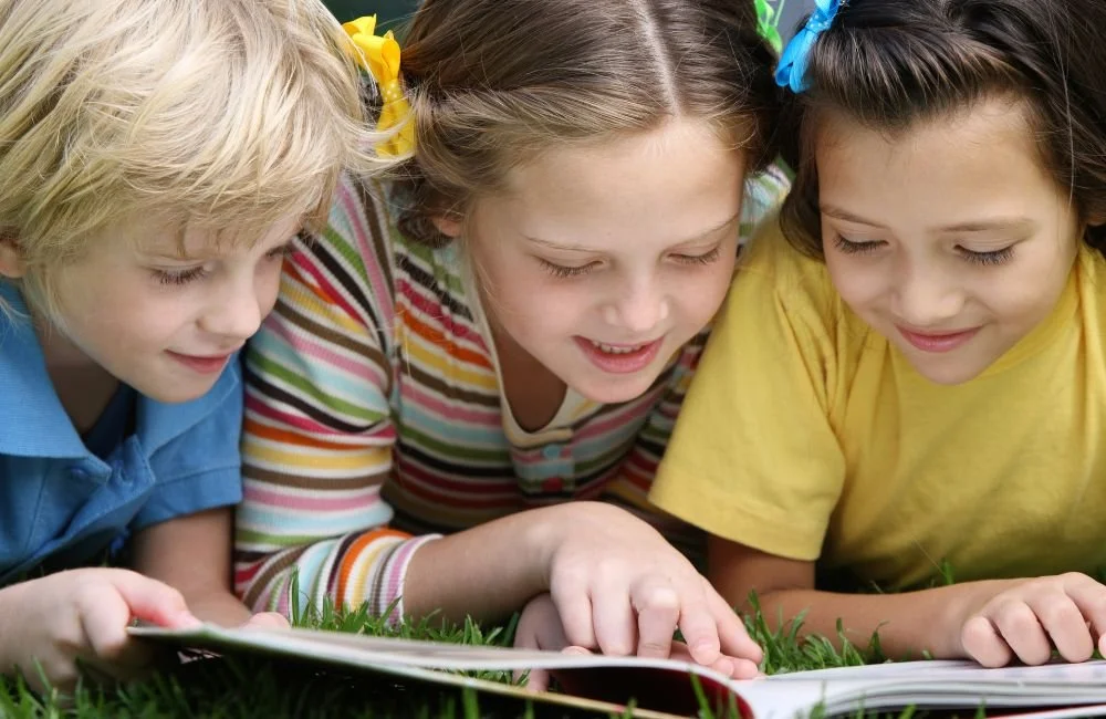 Three young kids looking closely at a book outside in the grass | Orange Seeds Montessori Centre
