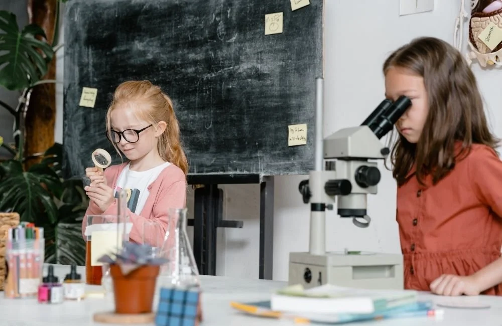 A little girl looking through a magnifying glass while another girl looks through a microscope in a science classroom