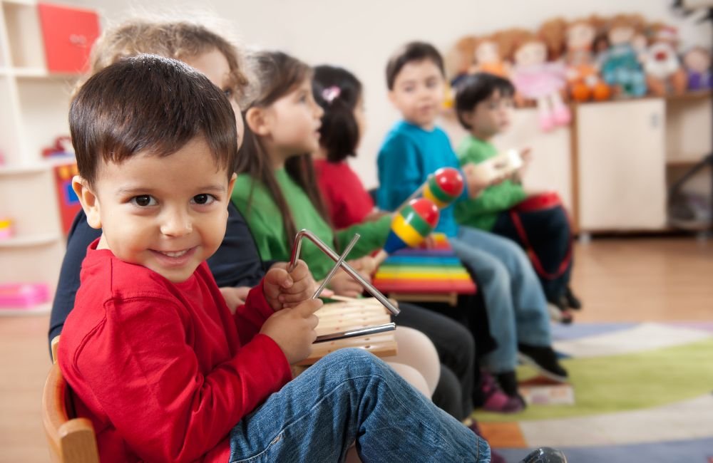 A little boy smiling in a lineup of preschoolers playing instruments in a Montessori classroom