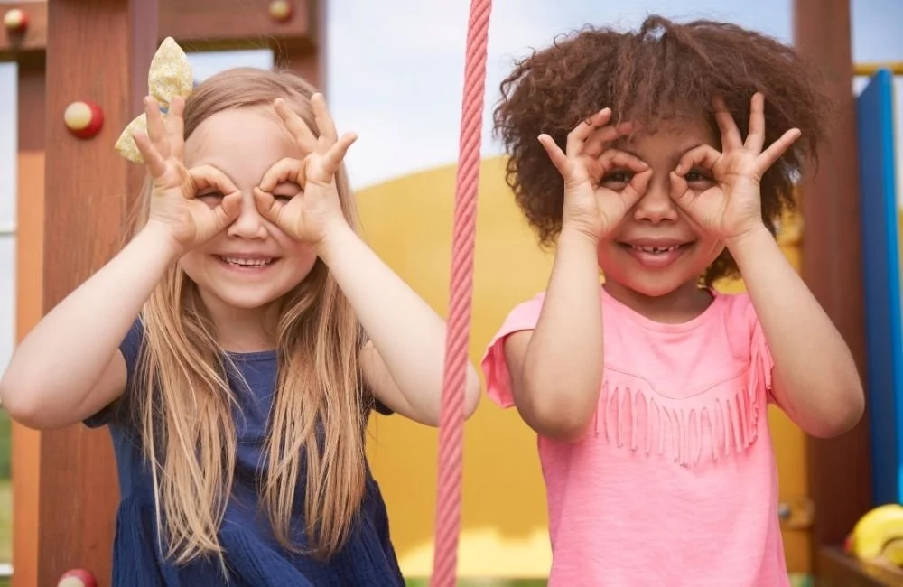 2 little girls smiling at the park with their fingers forming a mask over their eyes | Orange Seeds Montessori Centre