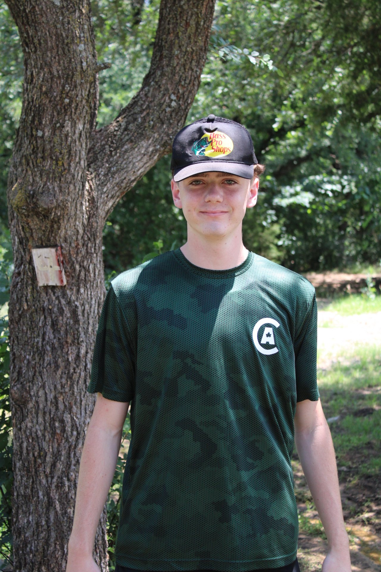 A young man stands outdoors in front of a tree, wearing a black cap with a Fishing Pro Shop logo and a dark green camouflage-patterned shirt.