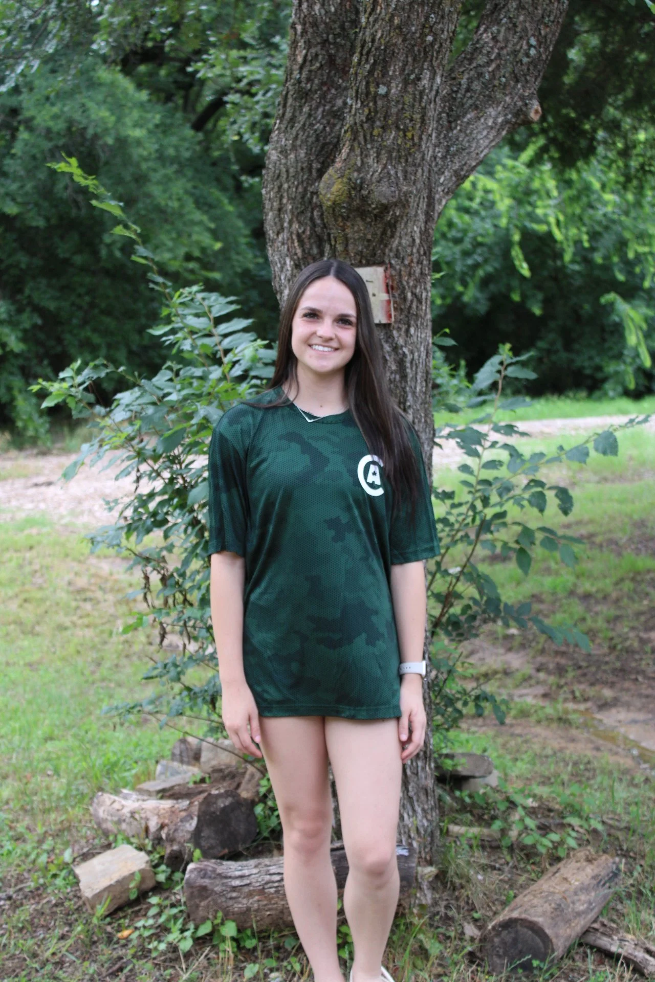 A young woman with long brown hair wearing a dark green sports jersey standing outdoors in front of a large tree and green foliage.