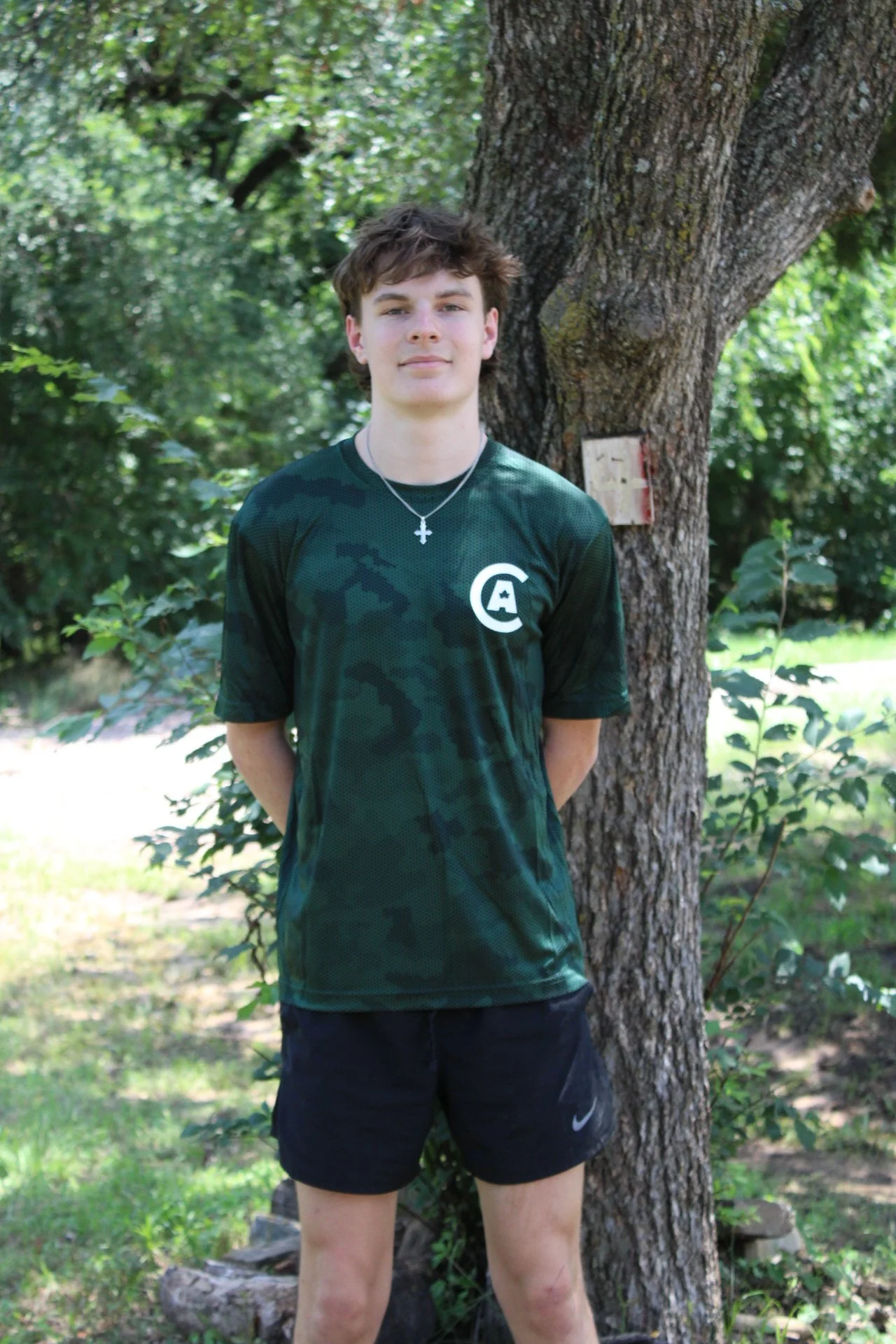 A young man with short brown hair standing outdoors near a large tree in a park or wooded area, wearing a green camouflage-patterned T-shirt with a white logo on the chest, black Nike shorts, a silver cross necklace, and looking at the camera.