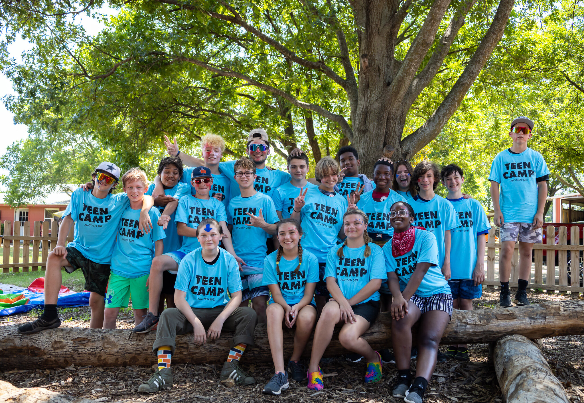 A group of children and teenagers wearing turquoise t-shirts that say "Teen Camp" gathered outdoors under a large tree with green leaves, smiling and posing for the photo.