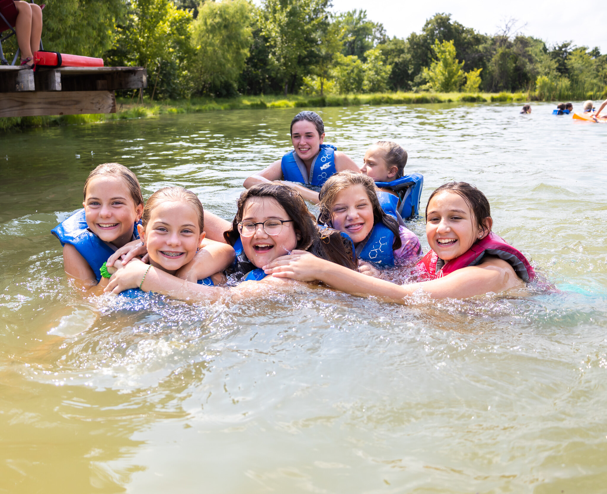 Group of children swimming and enjoying water at a lake, wearing life jackets, with a dock and lush trees in the background.