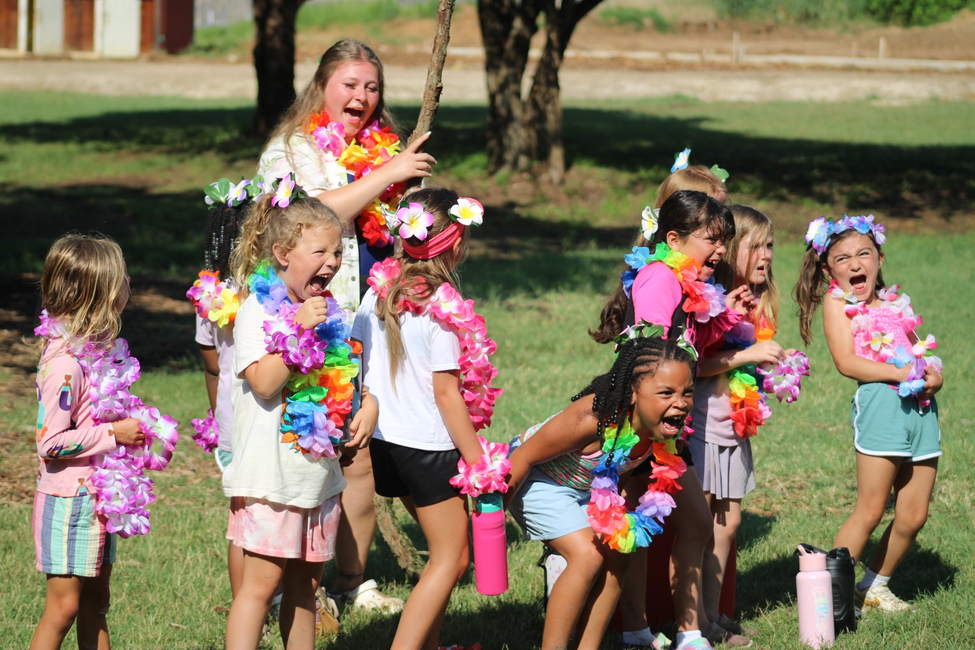 A group of children celebrating outdoors with leis, colorful accessories, and cheerful expressions, participating in a fun activity.