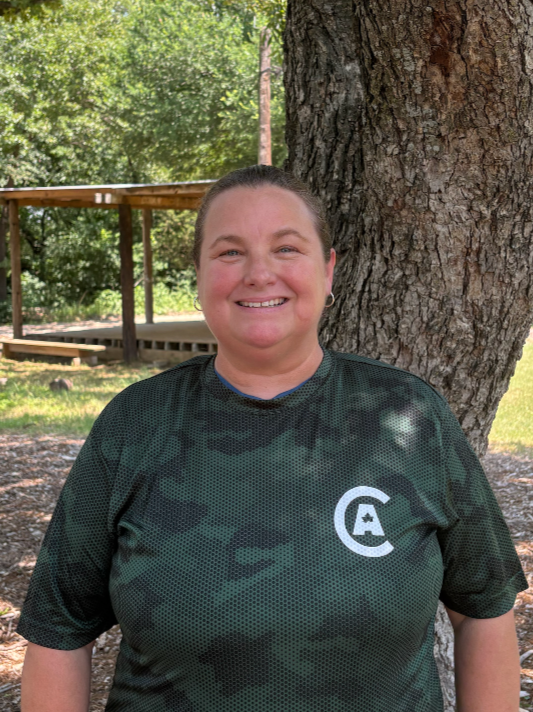 A smiling woman standing outdoors next to a large tree, wearing a dark green athletic shirt with a white logo, in a park setting with trees and a wooden structure in the background.