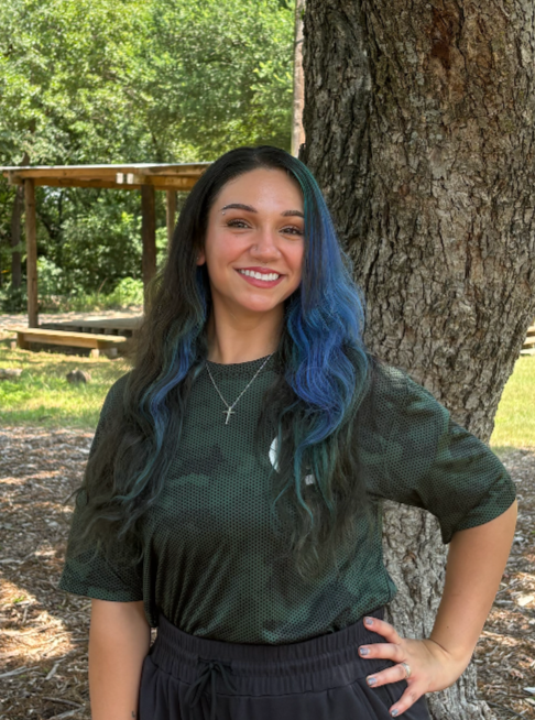 Young woman with long, wavy hair colored blue and green, smiling and standing outdoors next to a large tree with a pavilion and green trees in the background.