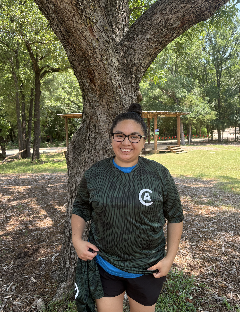 A smiling woman with glasses standing outdoors in front of a large tree, wearing a dark sports jersey and shorts, in a green park with trees and a small wooden structure in the background.