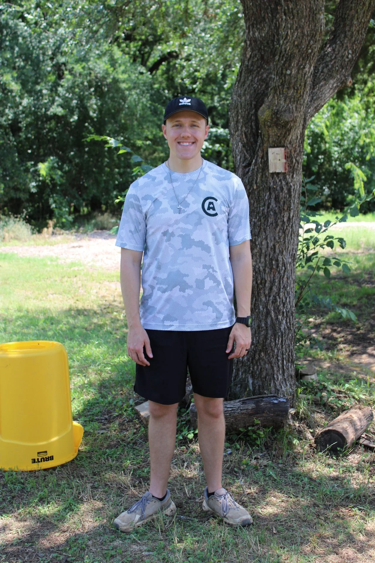A young man wearing a black Cap with Adidas logo, a camouflage patterned t-shirt, black shorts, and grey sneakers standing outdoors next to a large tree with textured bark and lush green foliage. There is a yellow plastic container on the ground nearby.