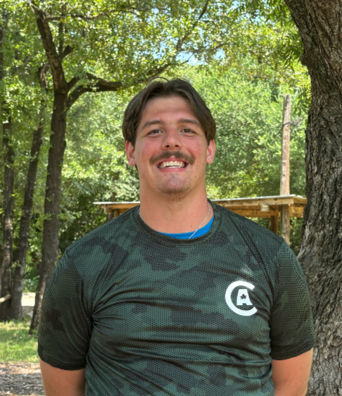 A young man with dark hair and a mustache smiling outdoors, wearing a black sports jersey with a logo, standing next to a tree in a wooded area.