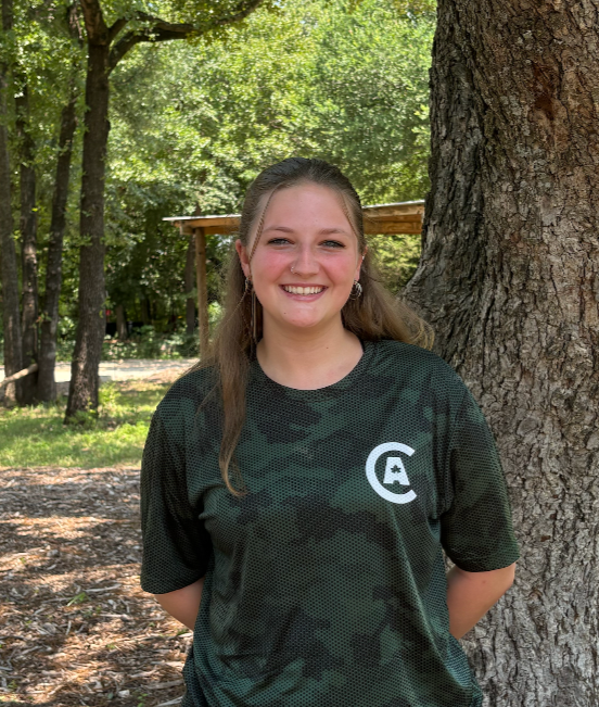 Smiling young woman with long brown hair standing outdoors beside a large tree in a park, wearing a black camo-patterned t-shirt with a white logo on the front.