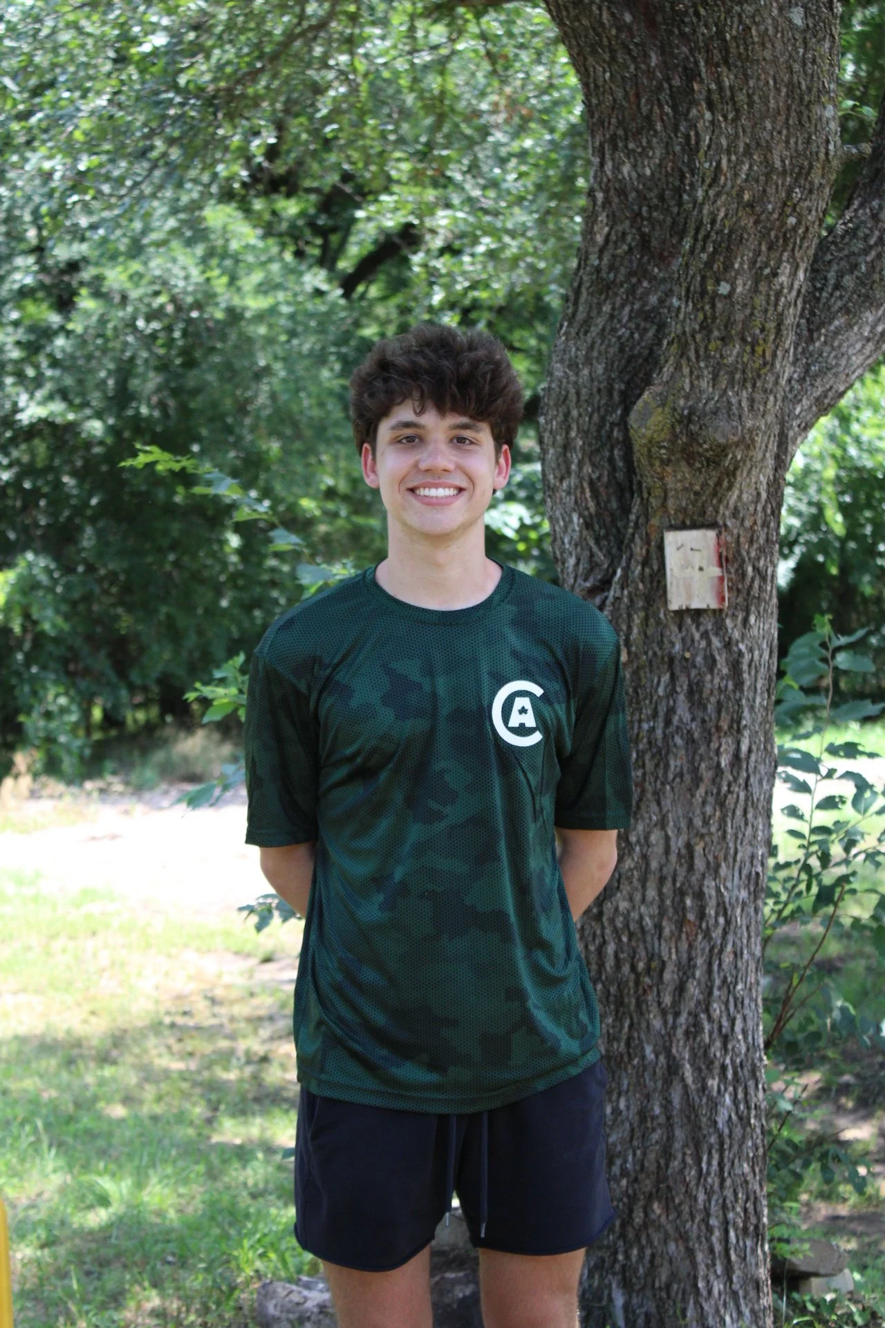 A young man with curly brown hair, smiling, stands outdoors next to a tree with green leaves in the background. He is wearing a dark green camouflage sports shirt and black shorts.