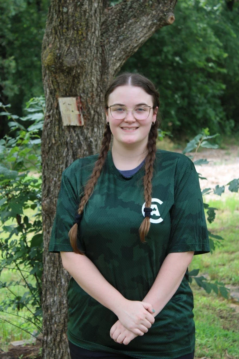 A young woman with glasses and braided hair smiling outdoors, standing in front of a tree with a small wood sign on it, surrounded by green foliage.