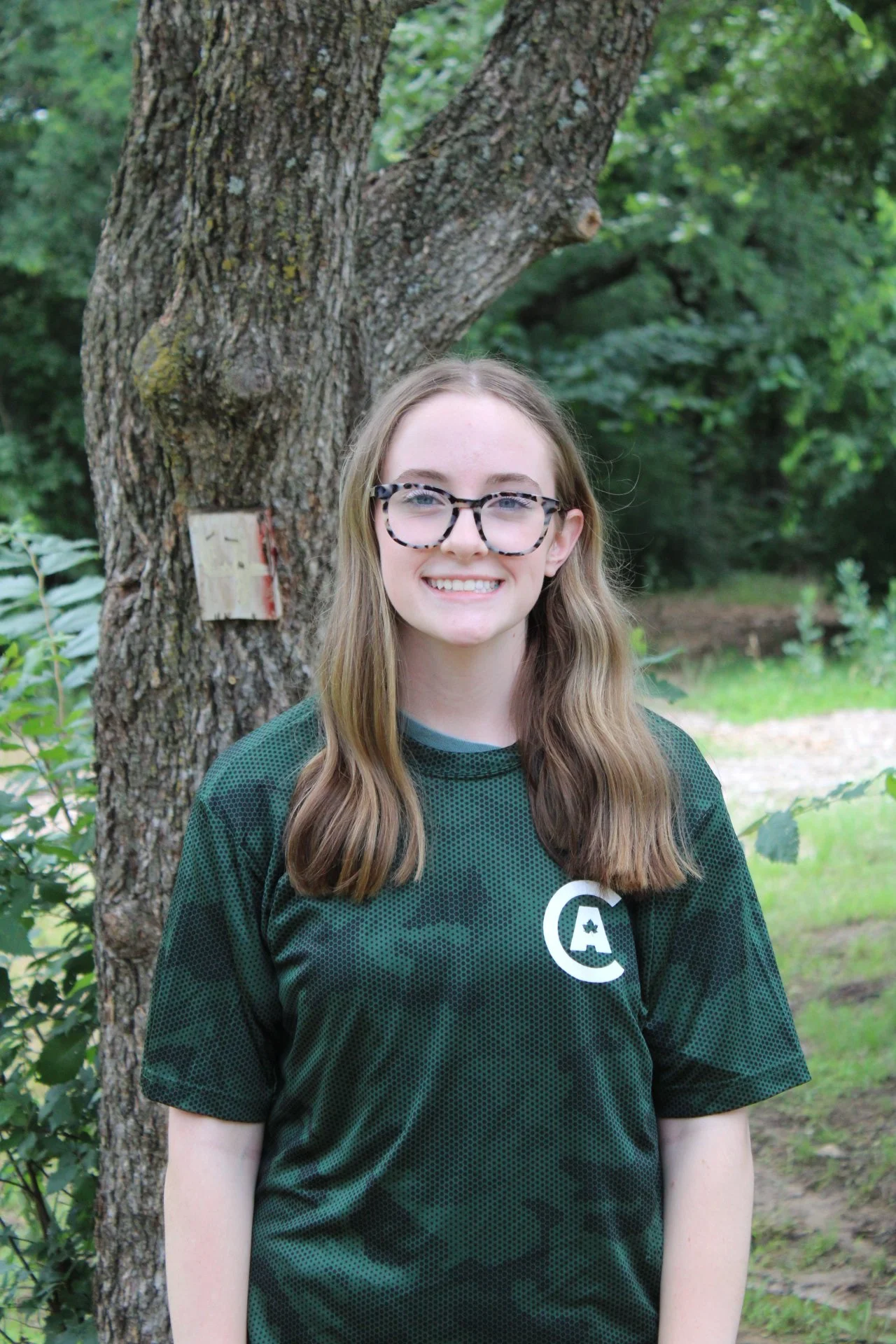 Young woman with long light brown hair, wearing glasses with a black and white pattern, smiling, standing outdoors in front of a tree with a brown trunk and green leaves, wearing a dark green sports jersey with a white logo.
