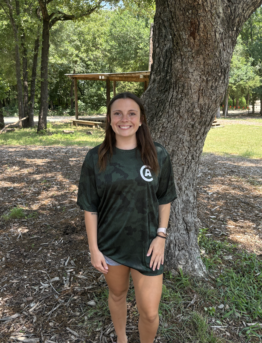 A young woman with brown hair smiling, standing outdoors in front of a large tree, wearing a black sports jersey with a white logo on it, and looking at the camera.