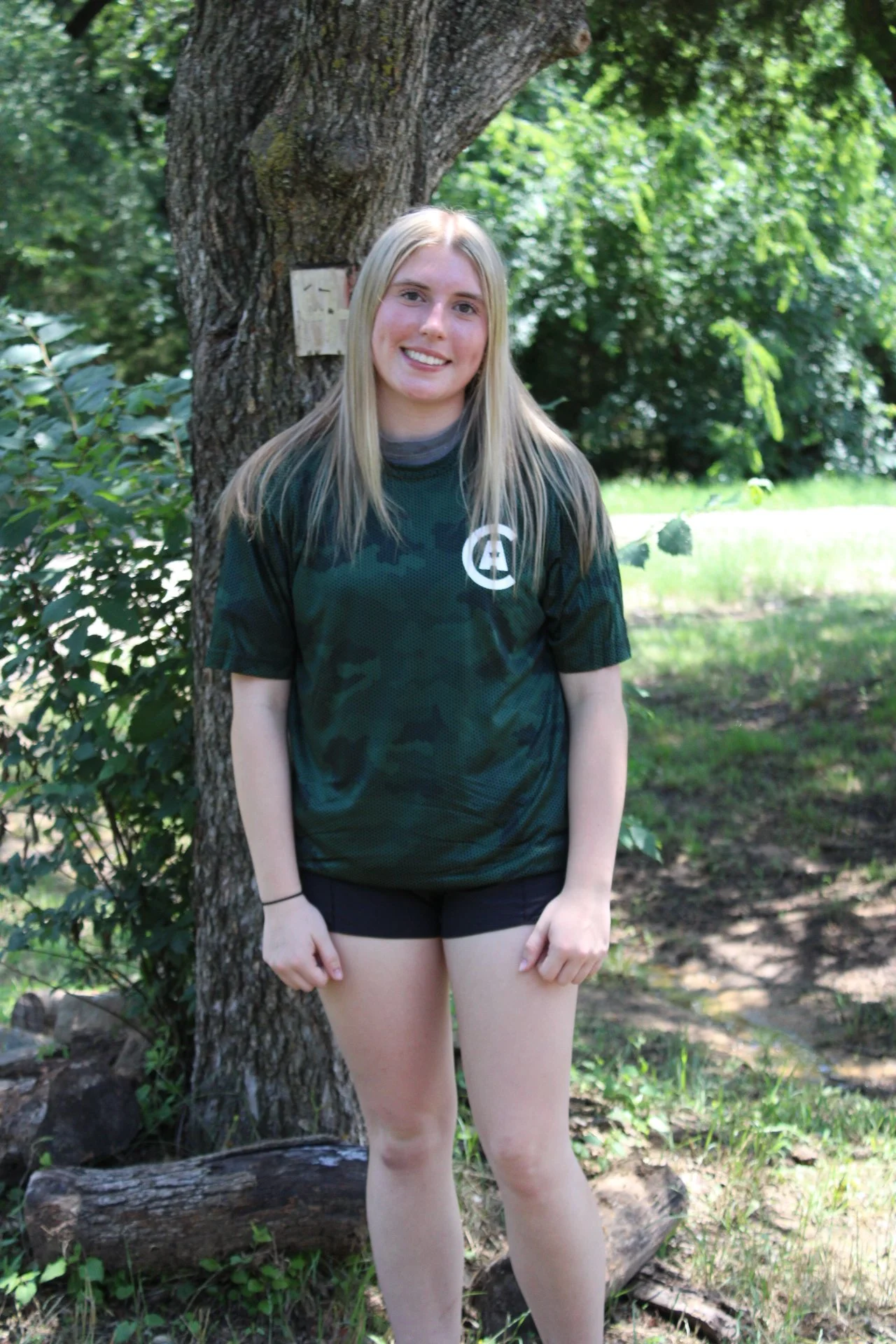 A young woman with long blonde hair standing outdoors in front of a tree, smiling, wearing a black camo sports shirt and black shorts.