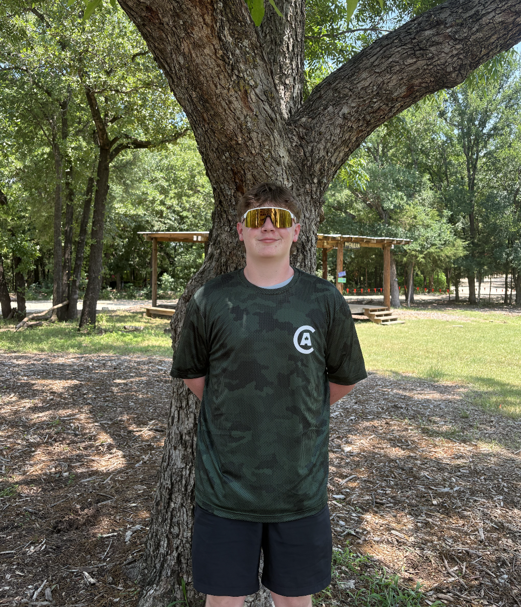 Camp Anothen Summer Staff, stands outdoors with his back against a tree, wearing reflective sunglasses, a black camouflage sports shirt with a white logo on the front, and black shorts, in a wooded park setting.