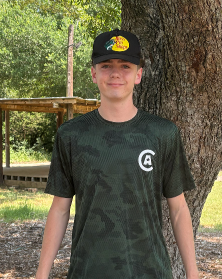 Young boy with a baseball cap and dark green camouflage T-shirt standing outdoors next to a tree.