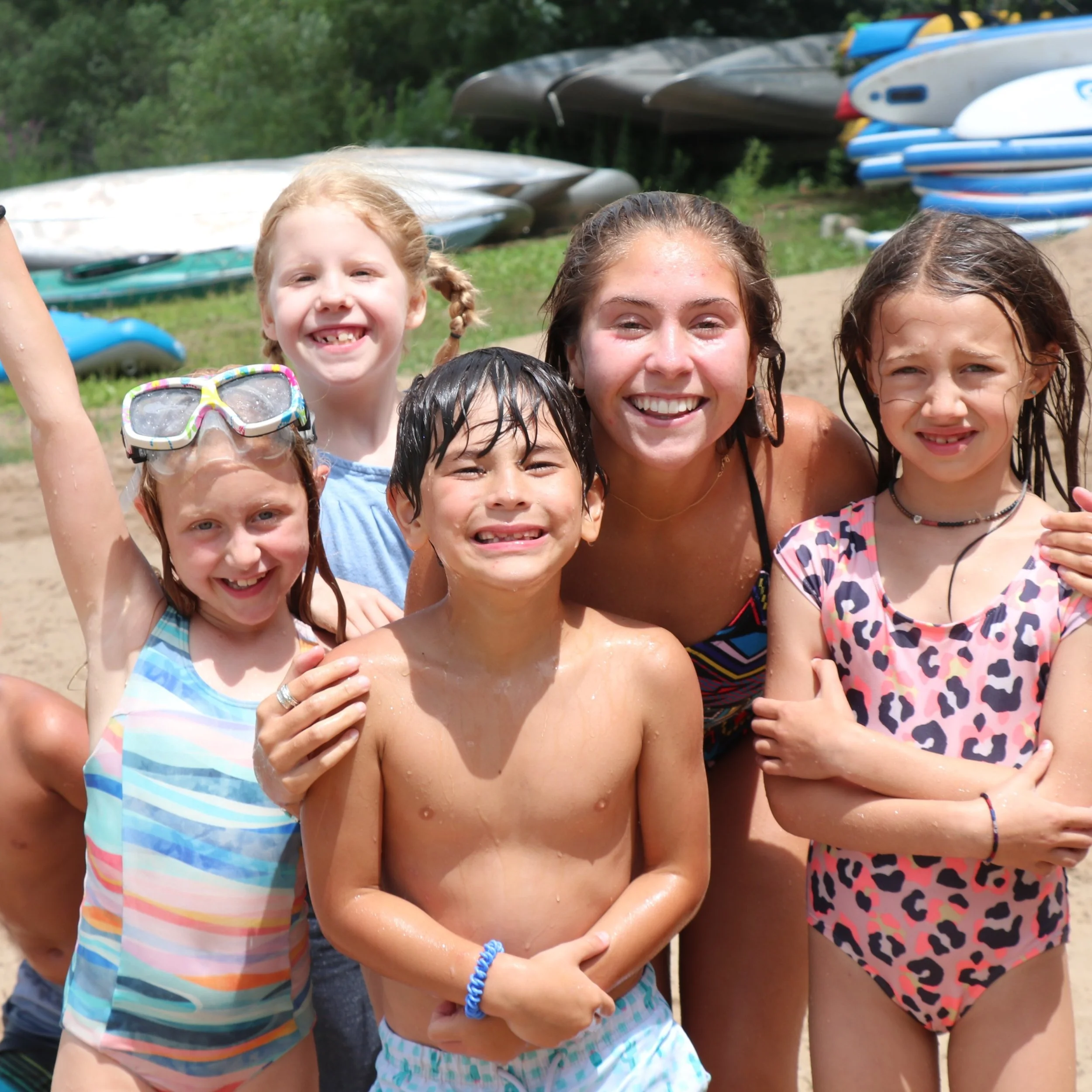 A group of children and a woman smiling outdoors near a beach with surfboards in the background.
