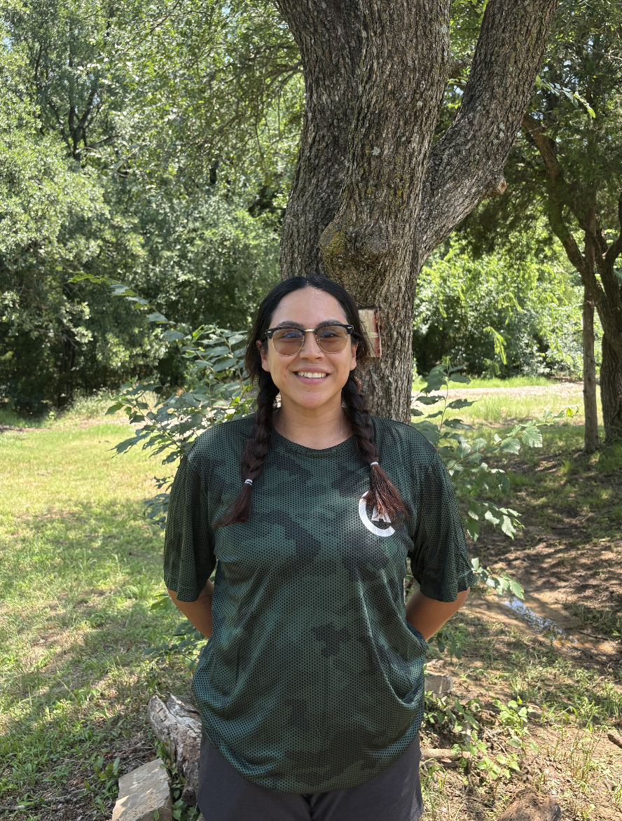 Young woman with braided hair, sunglasses, and a bright smile standing outdoors in front of a tree and greenery, wearing a dark sports jersey.