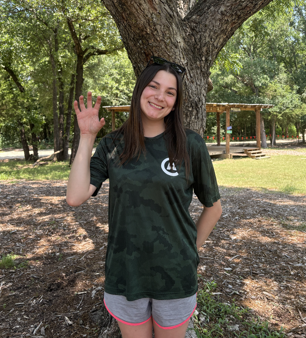 A young woman with long dark hair, smiling and waving, standing outdoors at a park next to a large tree. She is wearing sunglasses on her head, a dark green T-shirt, and gray shorts with pink trim.