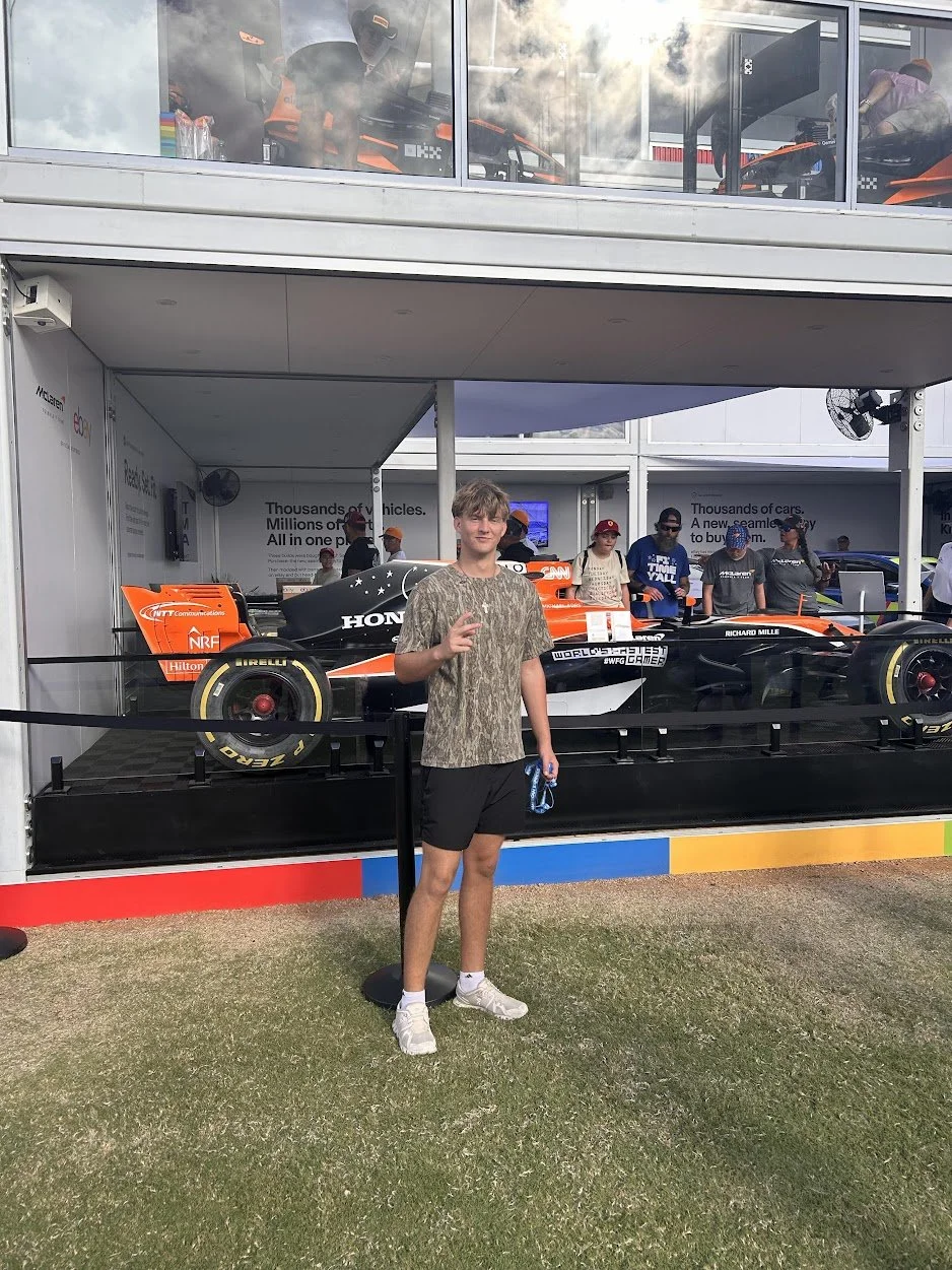 Young man with light brown hair and casual clothing making a peace sign in front of a racing car display at an event.