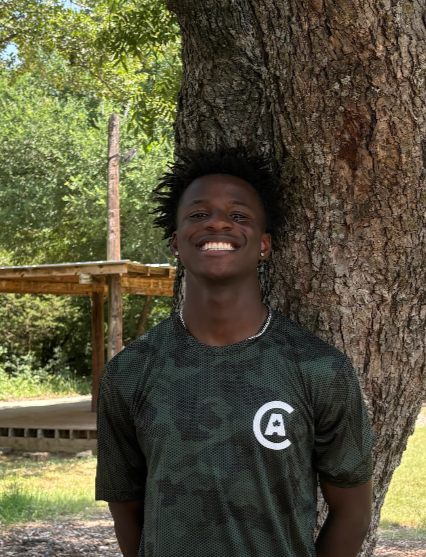 Young man with short curly hair smiling and standing against a large tree outdoors.