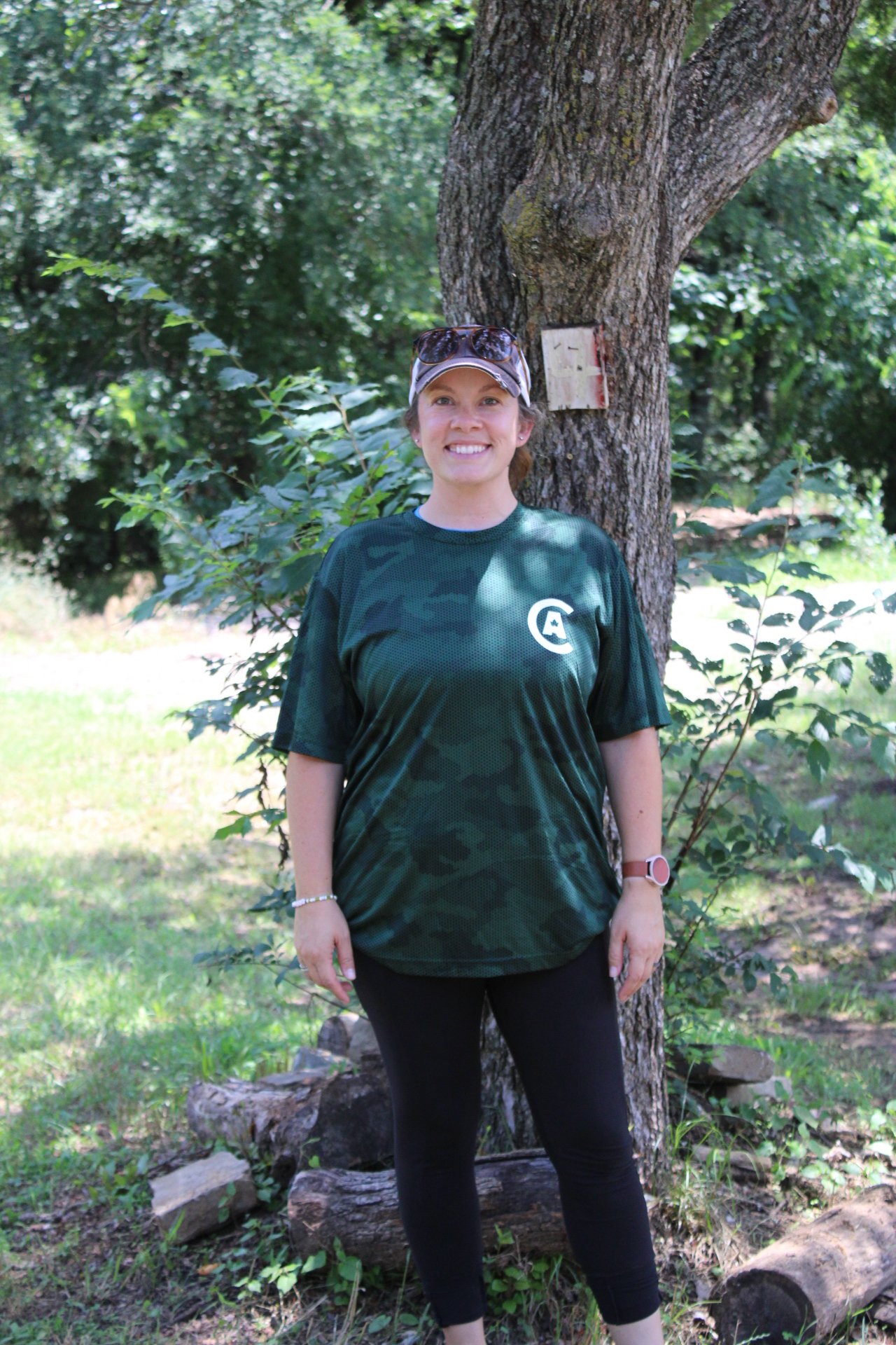 A woman standing outdoors in a wooded area with trees and greenery, smiling, wearing a camouflage-patterned shirt, black pants, a watch, bracelets, and sunglasses on her head, with a tree and a small sign behind her.