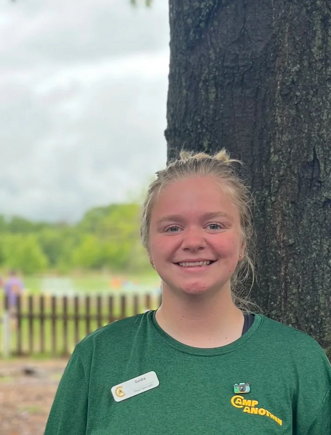 A smiling young girl with blonde hair and earrings, standing outdoors near a large tree with a wooden fence and green landscape in the background. She is wearing a green Camp Ivanhoe t-shirt and a name tag.