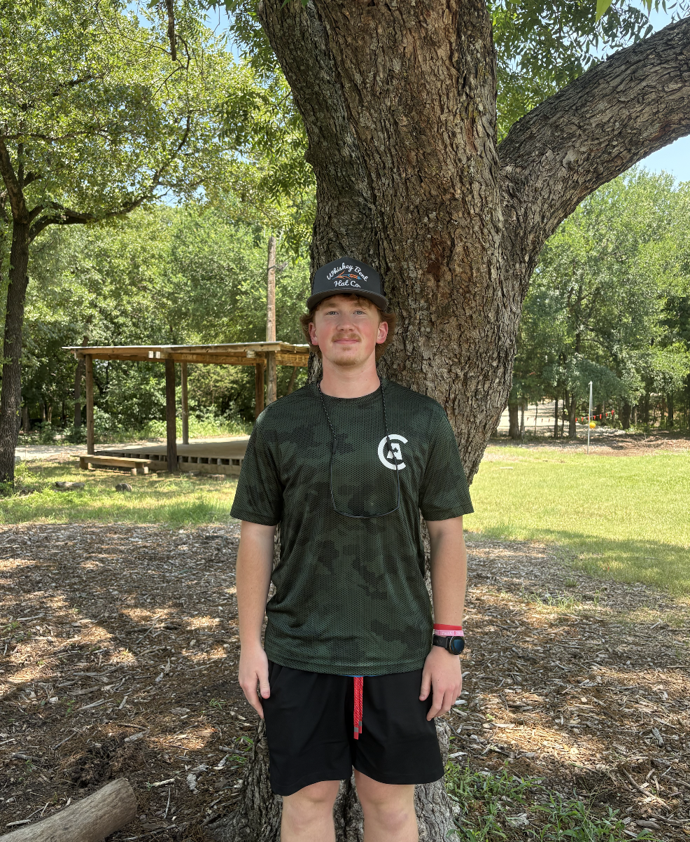 A young man standing outdoors next to a large tree with green leaves, wearing a dark green t-shirt, black shorts, a black cap, and a watch, with a wooden stage and green grassy area in the background.