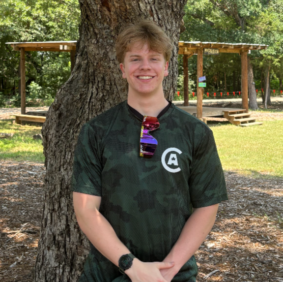A young man with blonde hair and a big smile, wearing a black camouflage-patterned shirt with a logo on the chest, sunglasses hanging from his collar, standing outdoors next to a large tree with a park or playground in the background.