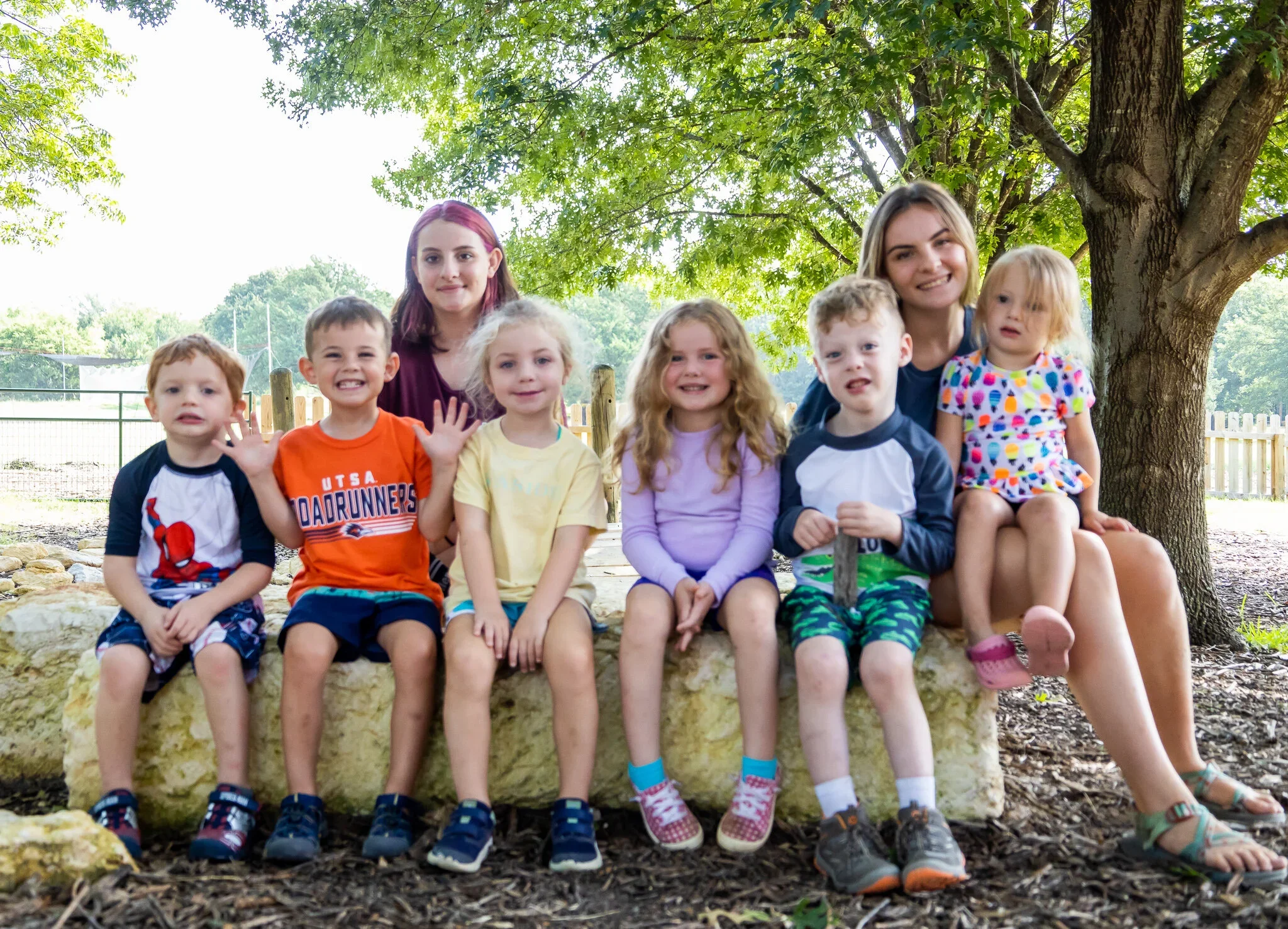 Group of young children and two adult women sitting outdoors under a tree, smiling and posing for the photo.