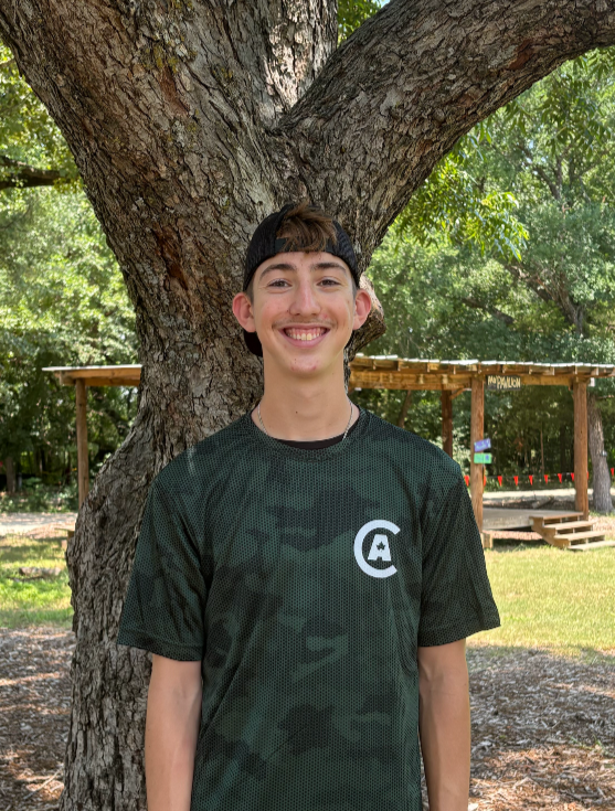 Smiling teenage boy wearing a black cap and camouflage sports shirt standing in front of a large tree outdoors with a wooden shelter in the background.