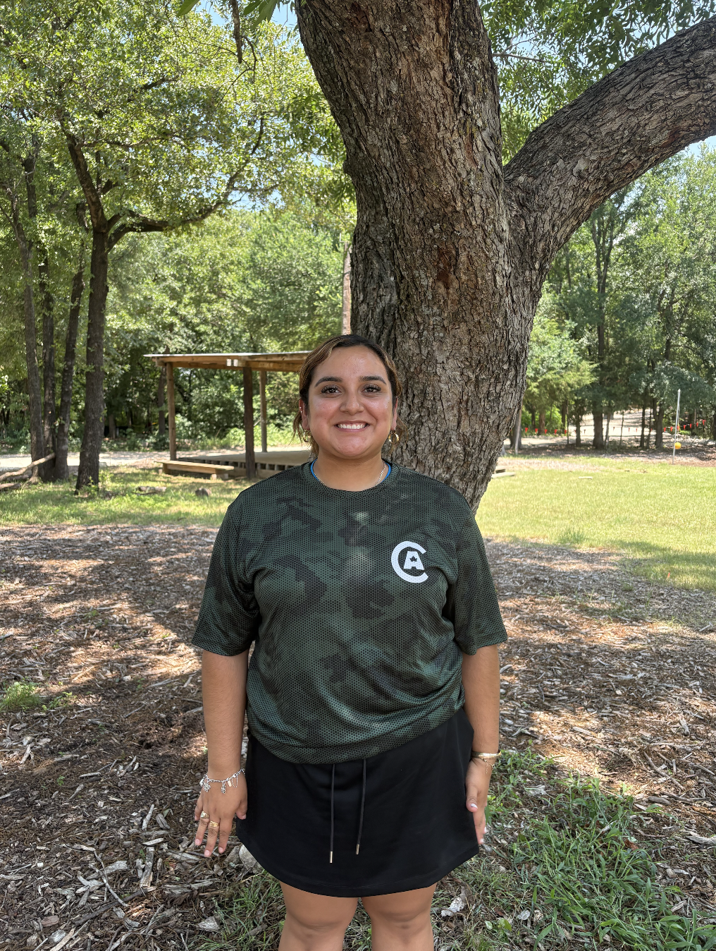 A woman standing outdoors near a large tree with lush green foliage, smiling at the camera. She is wearing a dark green camouflage-patterned shirt and a black skirt.