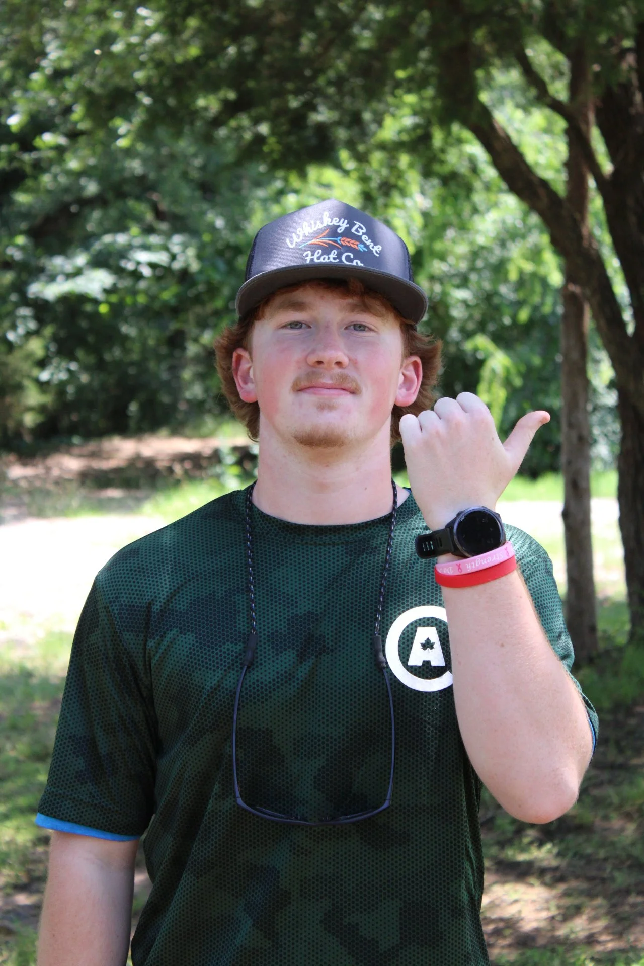 A young man with curly red hair and a slight mustache standing outdoors with trees in the background. He is wearing a black and green athletic shirt, a black digital wristwatch, a pink wristband, and a black cap with the text 'Lukey Bear Heat Co.' on it. He is showing a thumbs-up gesture with his right hand, which also has a necklace and sunglasses hanging around his neck.