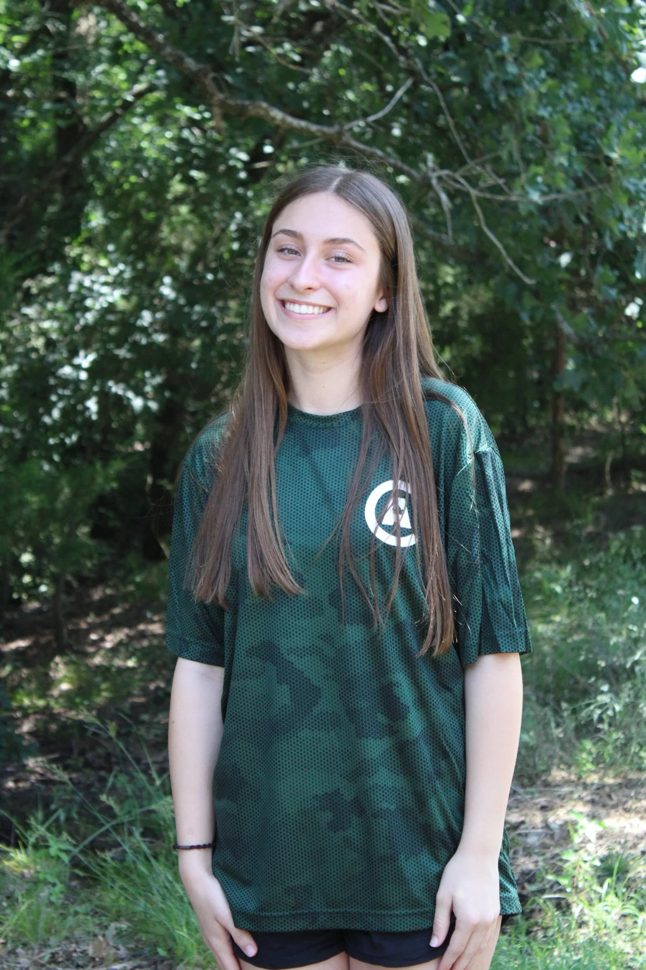 A young woman with long brown hair smiling outdoors with green trees in the background, wearing a dark green sports jersey with a white logo.
