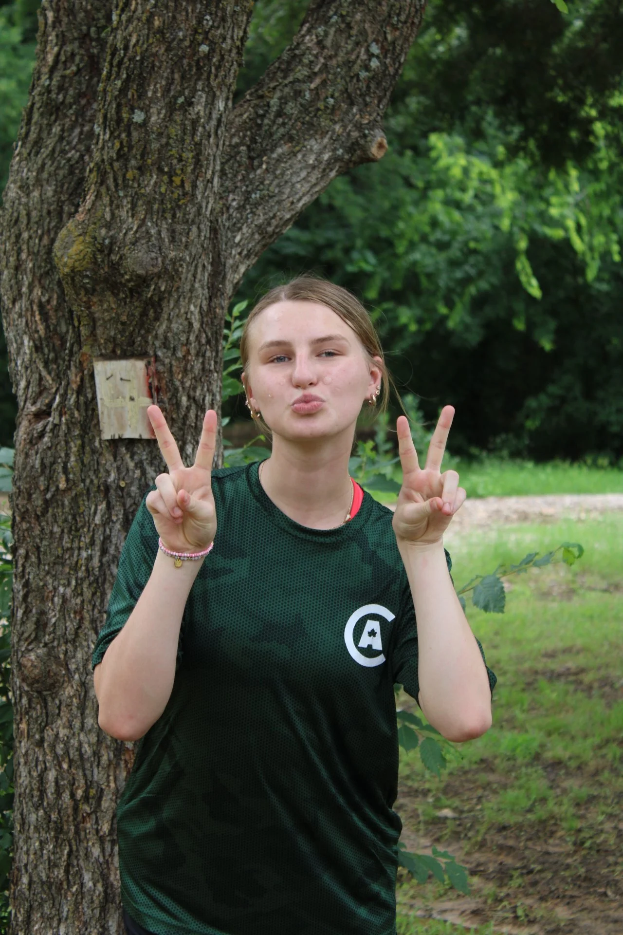 A girl standing outdoors next to a tree, making peace signs with her hands and posing for the camera. She has light skin, blond hair, and is wearing a dark green sports jersey.