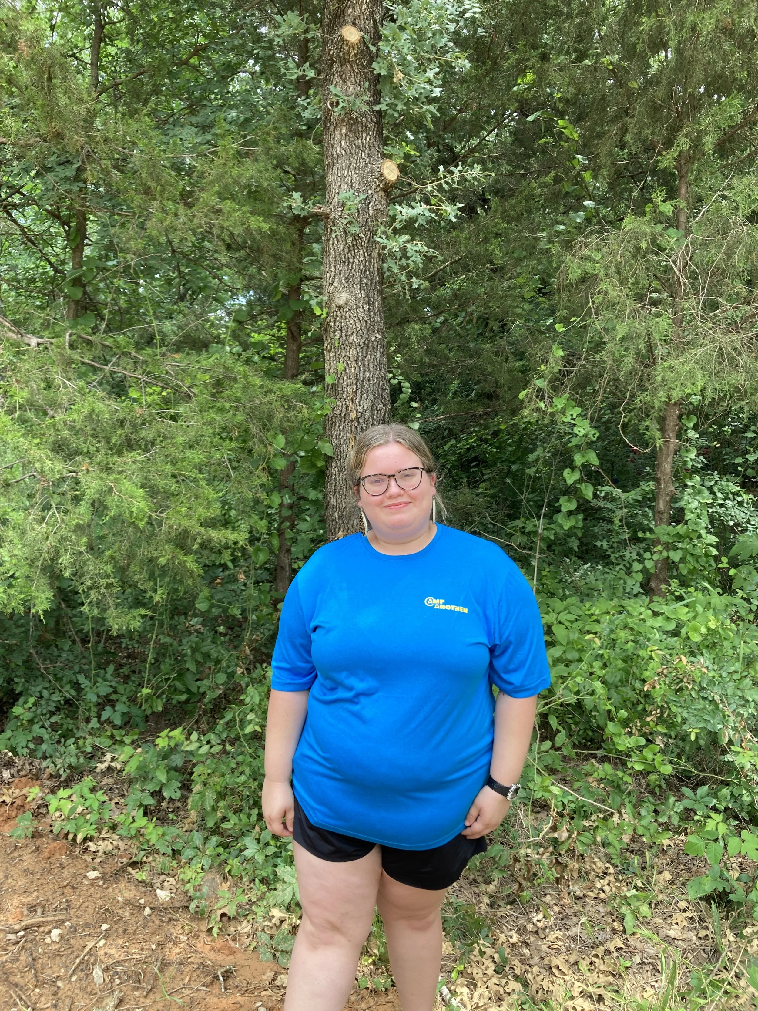 A woman wearing glasses, a blue t-shirt, and black shorts standing outdoors in front of lush green trees and plants.