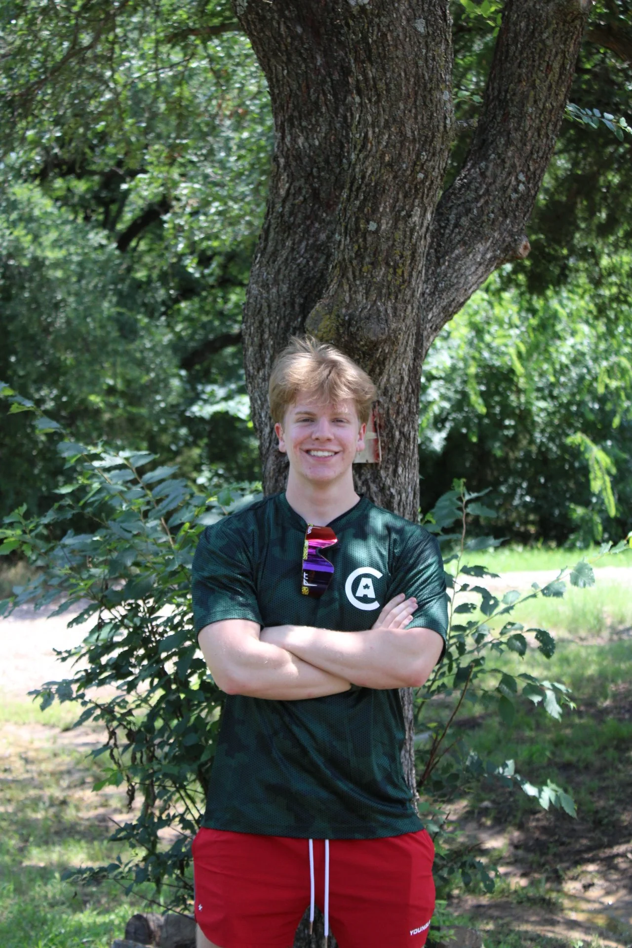 A young man with blond hair smiling and crossing his arms in front of a large tree in a park with green foliage.