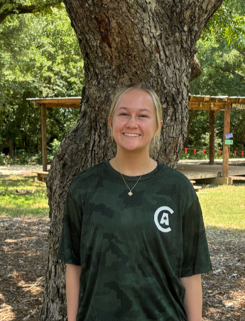 Young woman with blonde hair smiling, standing outdoors in front of a large tree. She is wearing a dark green sports jersey with a white letter A on it and a small necklace. Background includes a park with trees, a wooden structure, and a grassy area.