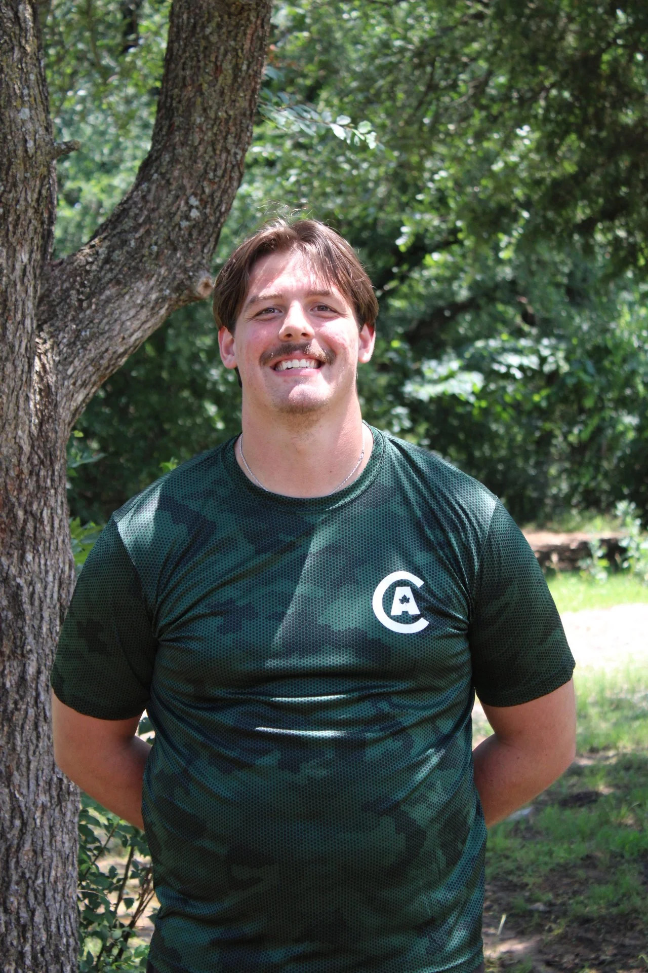 A smiling man with brown hair and a mustache stands outdoors near a tree, wearing a green camouflage-patterned athletic shirt with a white logo on the chest, background of green leafy trees and sunlight.