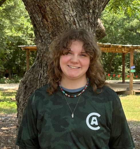 Smiling young woman with curly hair standing outdoors in front of a tree with a wooden shelter in the background.