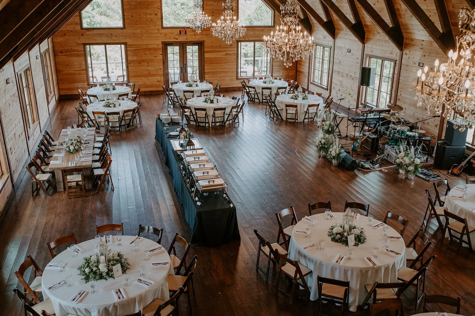 Rustic banquet hall with round dining tables, wooden chairs, chandeliers, and floral centerpieces; buffet table setup and a small stage with musical instruments.